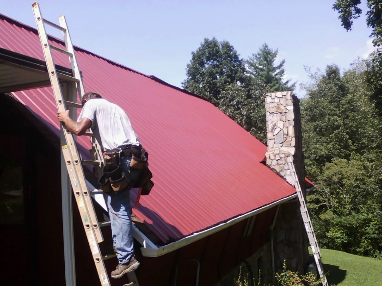 Roofer on a ladder beside a red metal roof and stone chimney — Soffit & Fascia Repair by Roofing Brothers in Waring